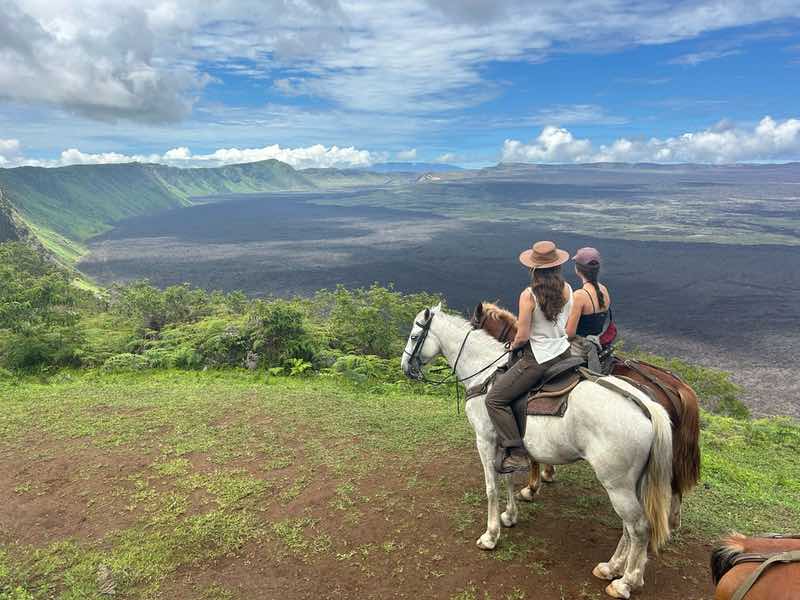 Caption: Riders pause above the vast Sierra Negra caldera, taking in the view toward the historic sulfur mines, Isabela Island, Galápagos.