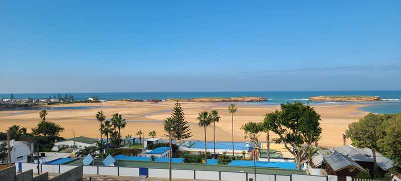 Wide view of Morocco's Atlantic coast with a broad sandy beach, palm trees, rocky islands and blue ocean.