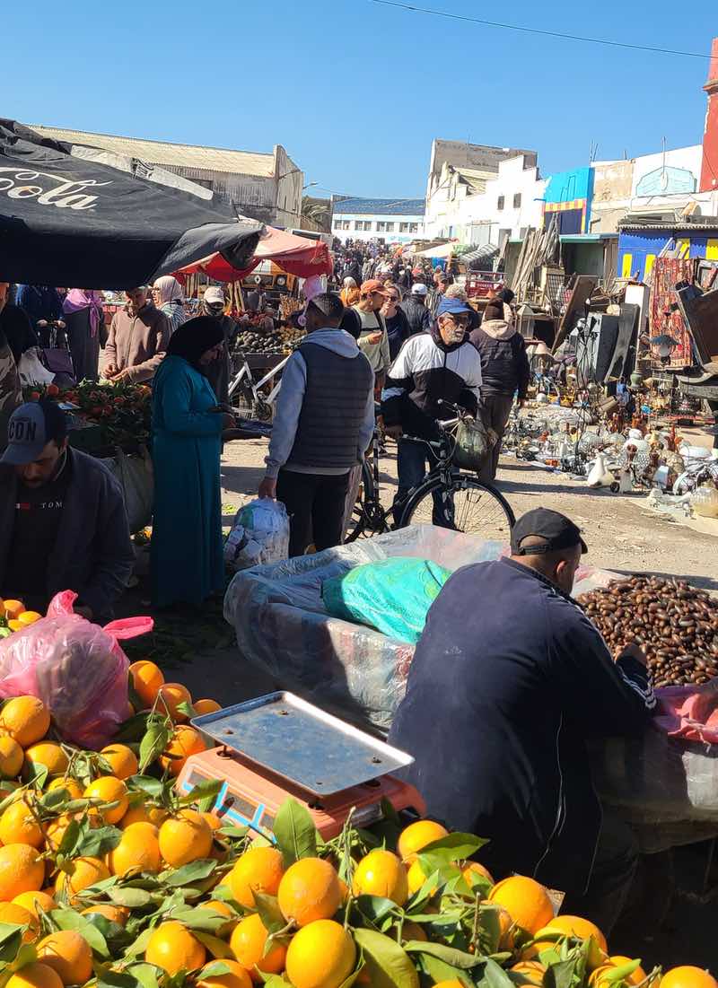 Fresh oranges in the foreground of a crowded outdoor souk in Morocco, with shoppers, vendors, produce stalls and bicycles filling the market.