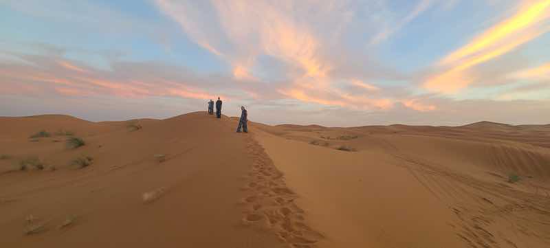 Travelers walking along a sand dune at sunset beneath pink clouds in the Sahara Desert in Morocco.